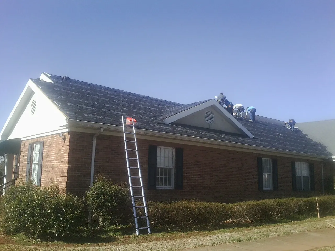 Crew tearing shingles off a brick ranch roof with a ladder set — Metal Roof Repair work by our crew in Norwood